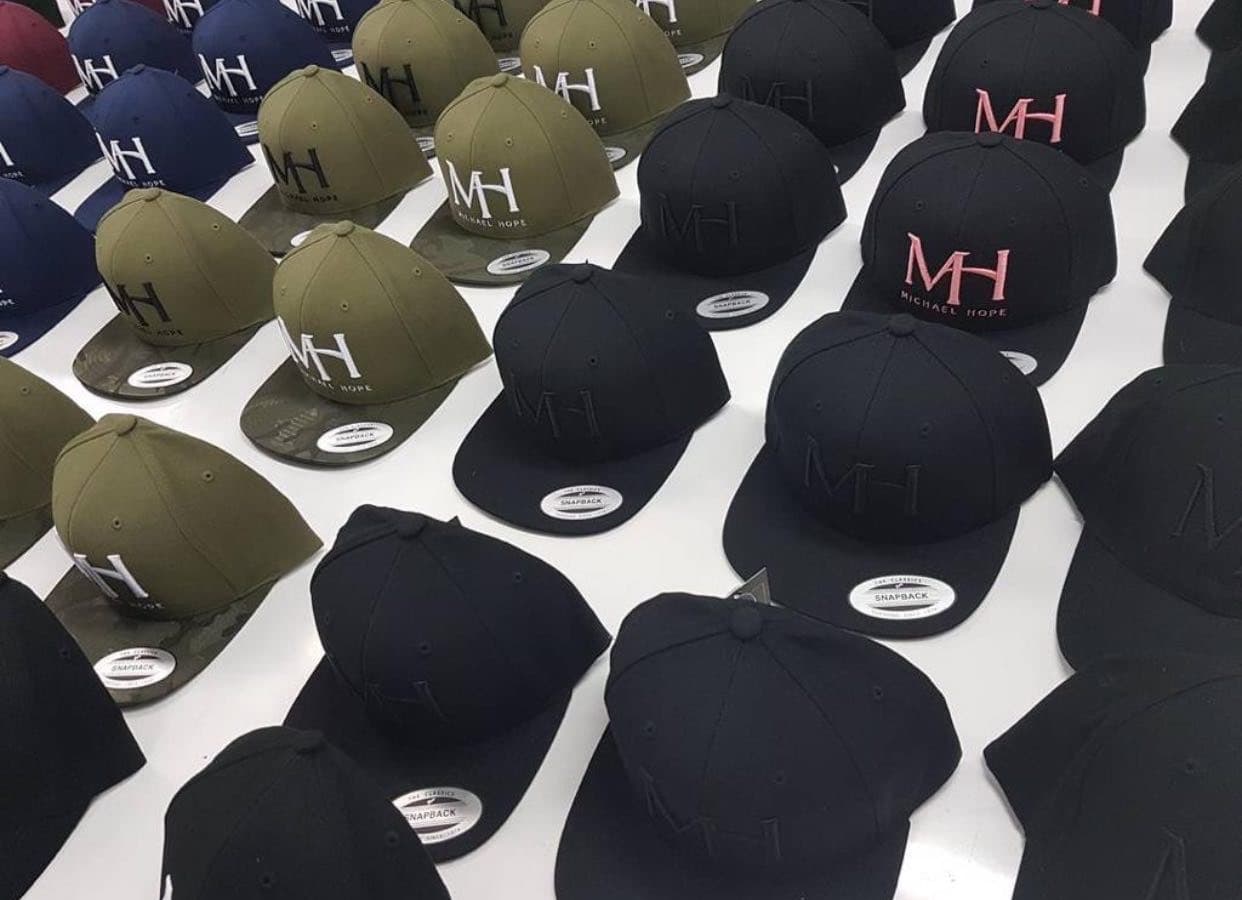 Rows of embroidered snapback caps in black, olive, navy and burgundy laid out on a white table
