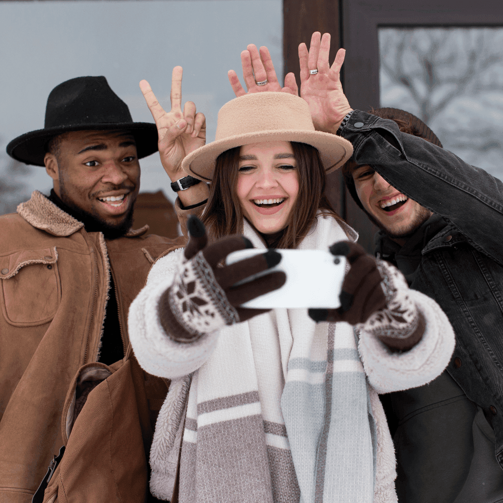 Three friends in winter coats, scarves and hats laughing and taking a selfie outdoors