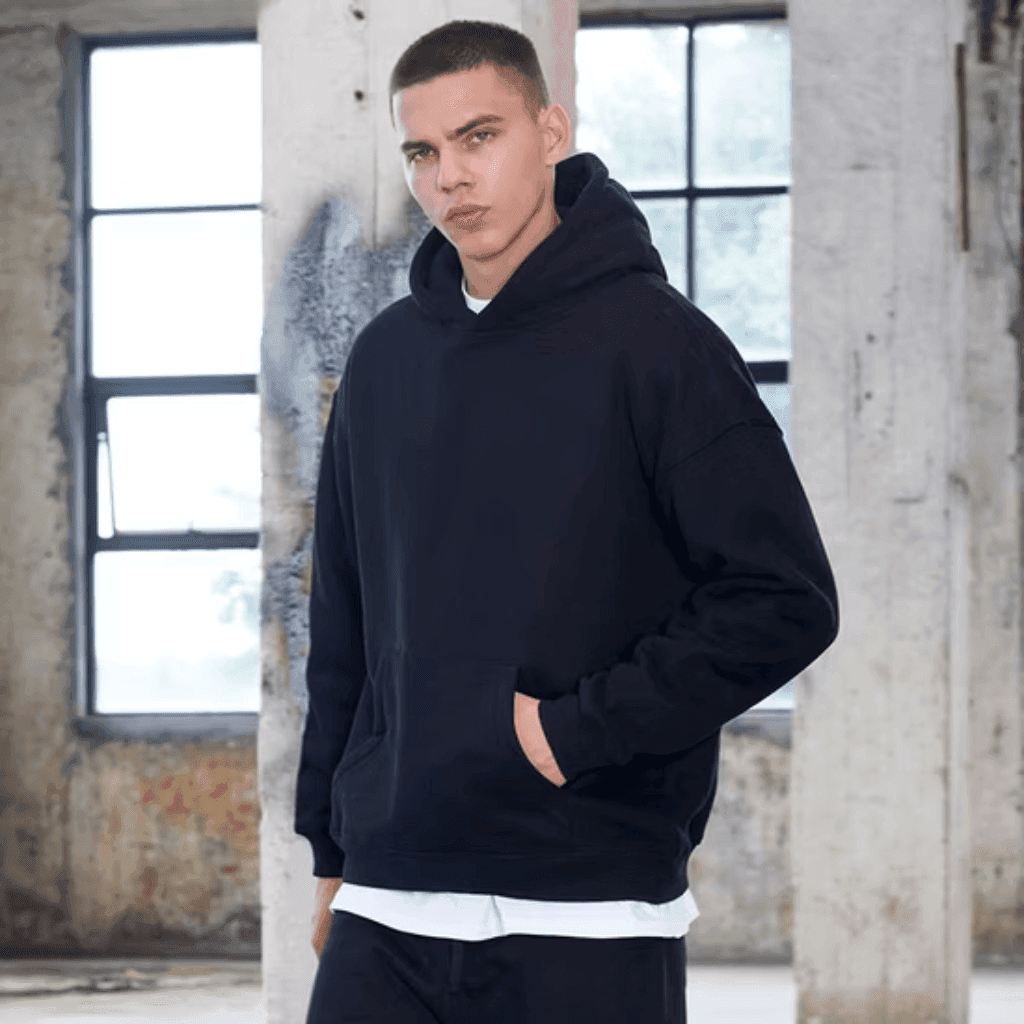 Young man in a black hoodie standing by a tall window in an industrial loft