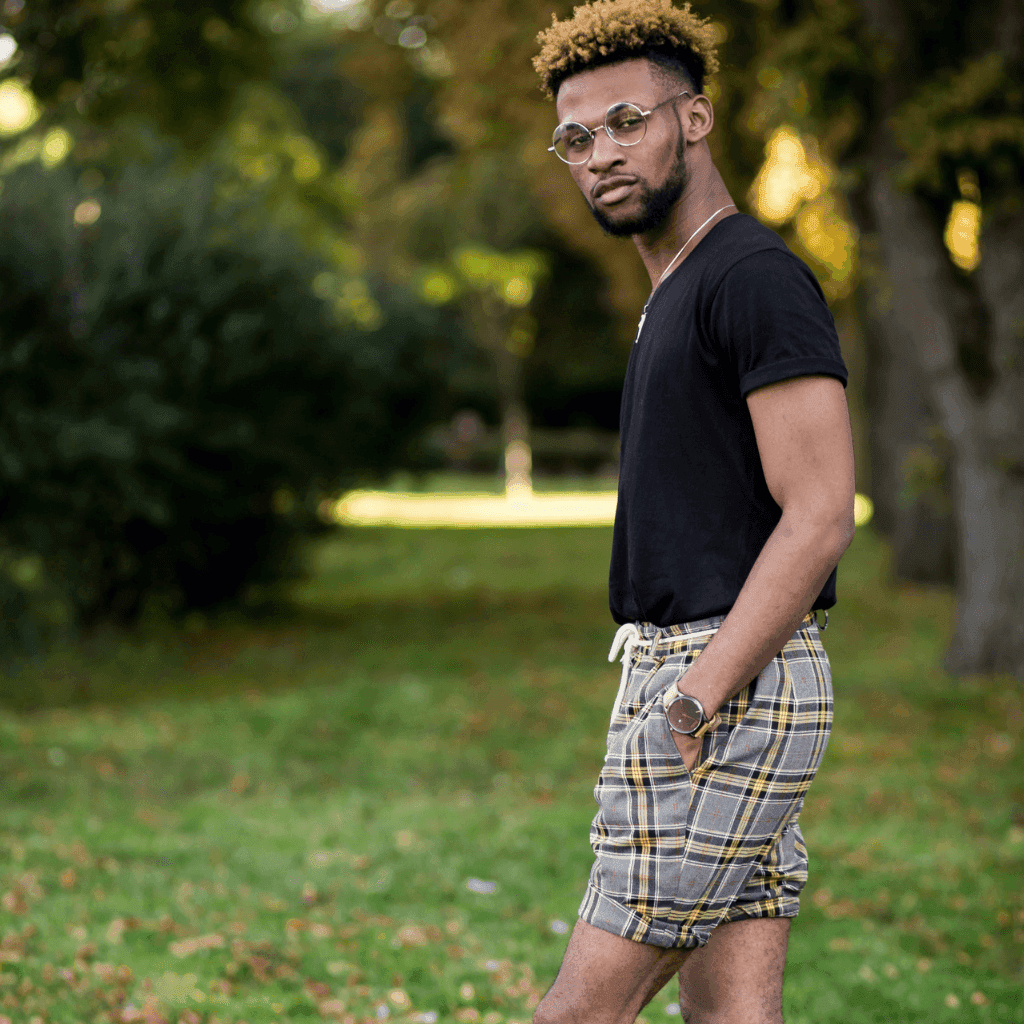 Man in round glasses, a black t-shirt and grey and yellow plaid shorts standing in a sunlit park