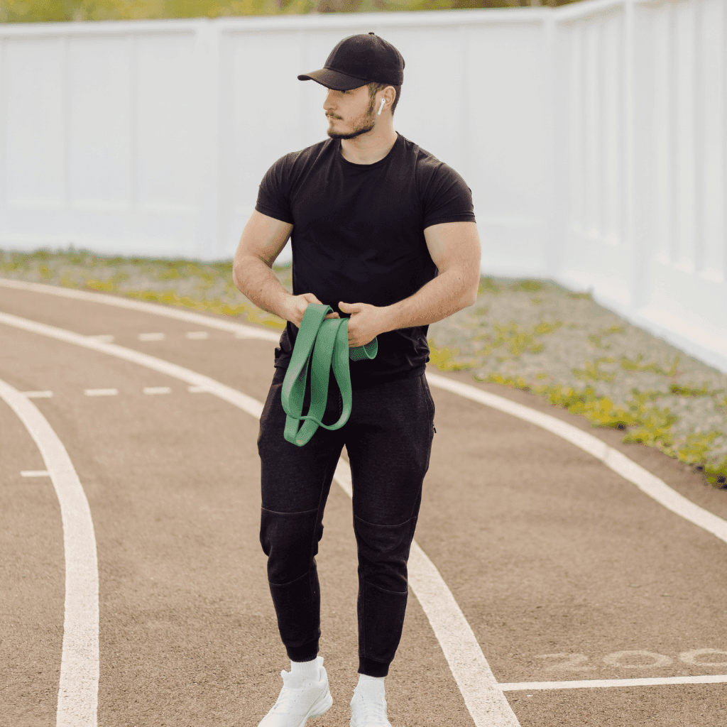 Man on an outdoor running track in a black t-shirt, black joggers and a black cap, holding a green resistance band
