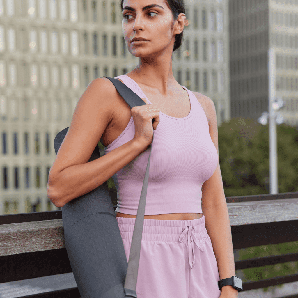Woman in a pink sports bra and matching shorts carrying a rolled yoga mat outdoors