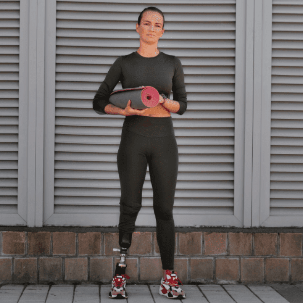 Woman with a prosthetic leg in an olive long-sleeve top and leggings, holding a rolled yoga mat in front of grey shutters