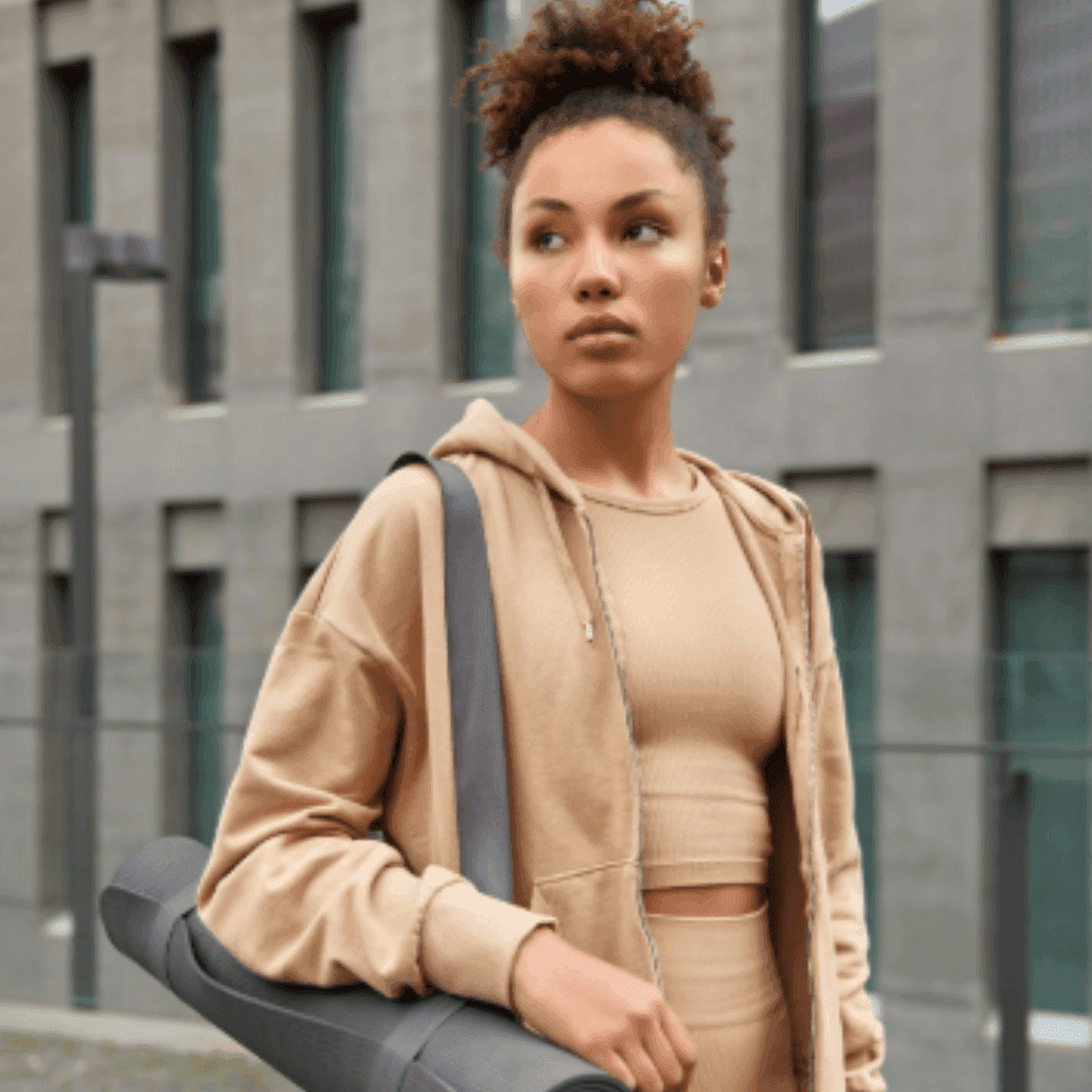 Woman in a beige zip hoodie and matching crop top with a shoulder bag, standing outside a modern building