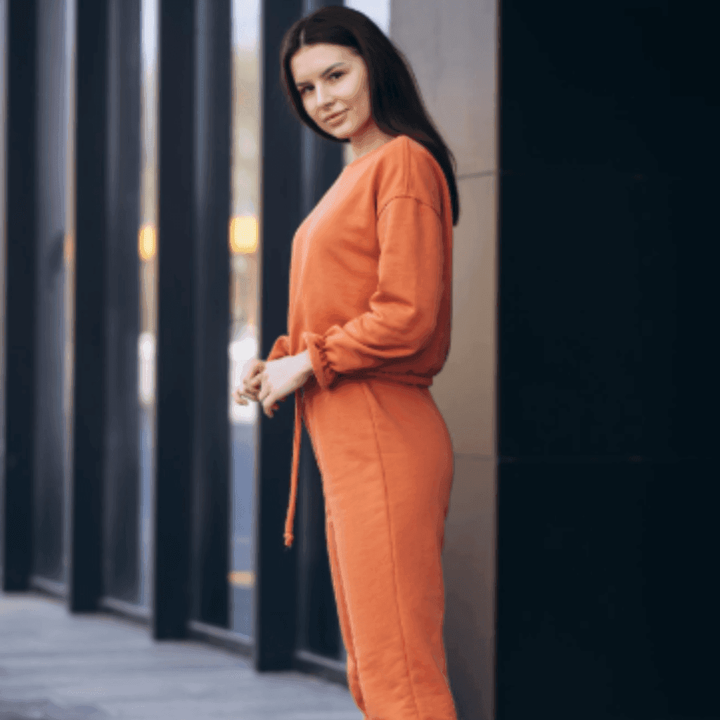 Woman in an orange zip-up top and matching trousers standing against a dark wall