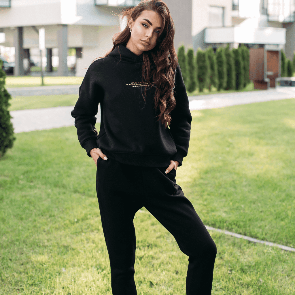Woman in a black hoodie and black joggers standing on a lawn in front of a building