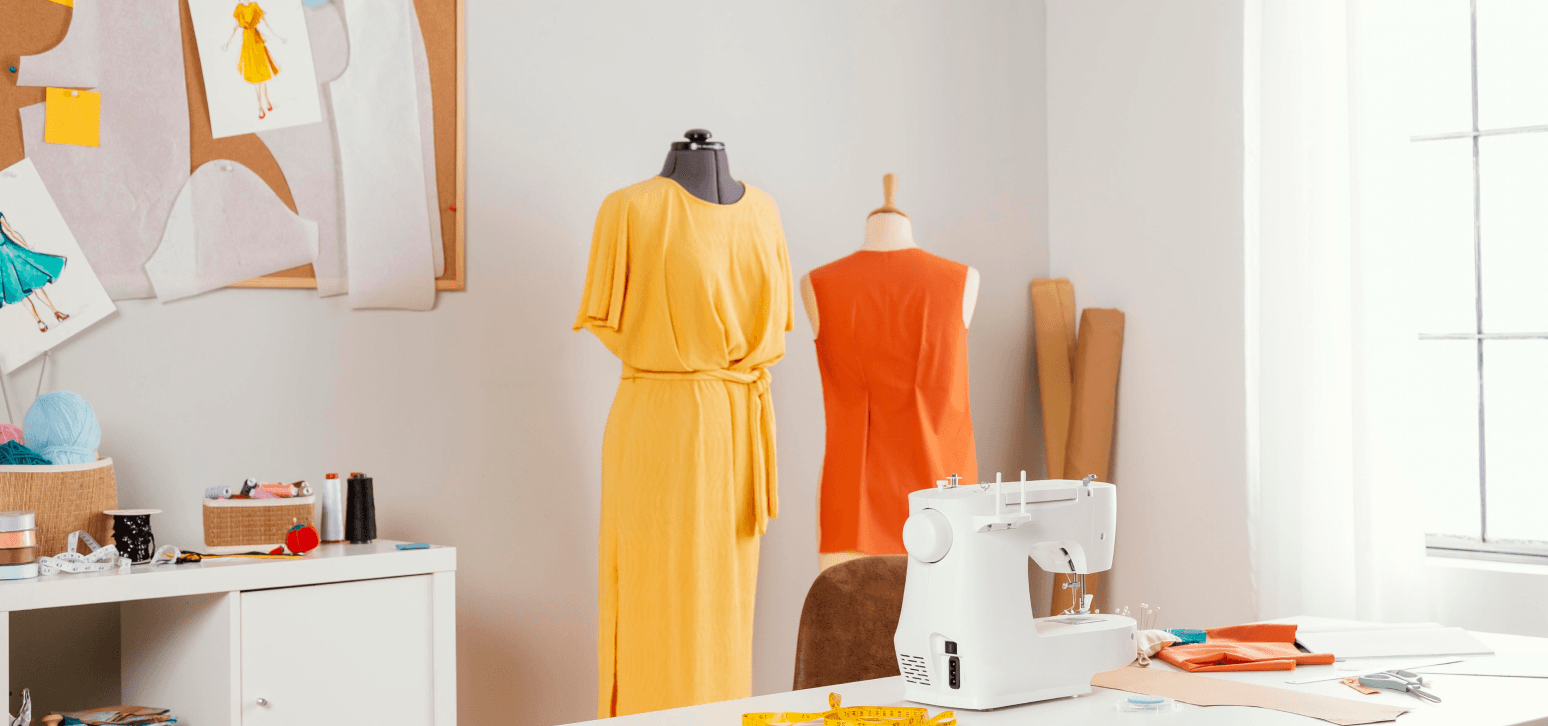 Design studio with a yellow dress and an orange top on mannequins beside a sewing machine